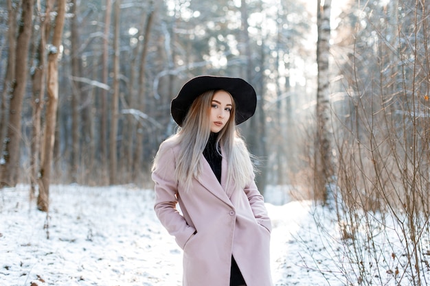 Nette schöne junge Frau mit blondem Haar in einem Vintage-schicken schwarzen Hut in einem rosa eleganten Mantel, der in einem Winterpark aufwirft. Attraktives stilvolles Mädchen.