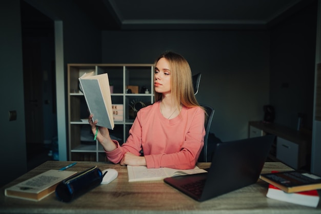 Nette Studentin, die zu Hause am Schreibtisch mit Laptop studiert und ein Buch mit ernstem Gesicht liest