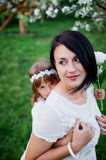 Nette Tochter und Mutter, die im blühenden Frühlingsgarten umarmen glückliche Frau und Kind, weiße Kleidung im Freien tragend, Frühlingssaison kommt. Muttertagsferienkonzept