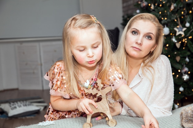 Nette Tochter und Mutter, die Weihnachten feiert. Eltern und kleines Kind, die sich in der Nähe des Weihnachtsbaums im Haus amüsieren, Feiertage, Feiern, Familien- und Menschenkonzept