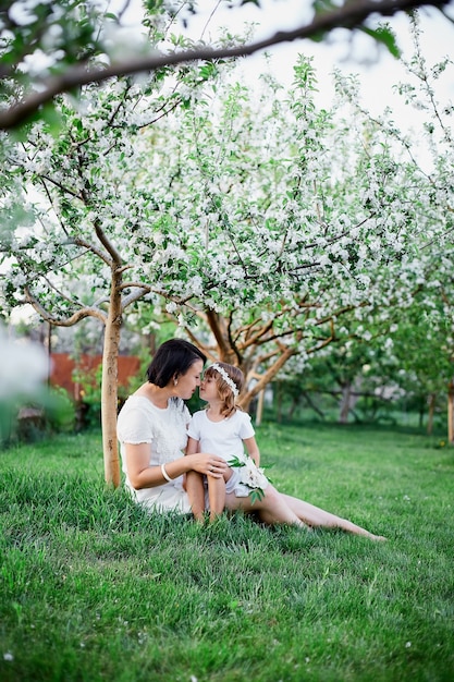 Nette Tochter und Mutter sitzen und umarmen im blühenden Frühlingsgarten Glückliche Frau und Kind, tragen weißes Kleid im Freien, Frühlingssaison kommt. Muttertagsferienkonzept