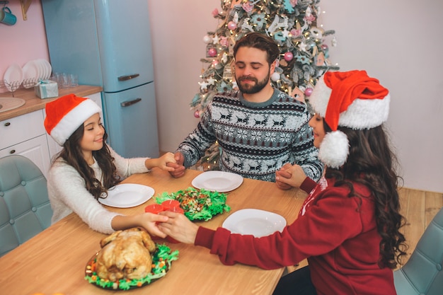 Nette und entzückende Familienmitglieder, die am Tisch sitzen und spielen. Sie halten sich an den Händen und halten die Augen geschlossen. Sie beten, bevor sie festliches Essen essen.