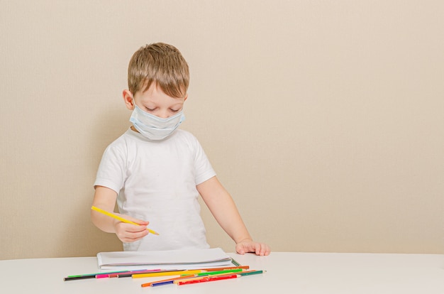 Netter 4 Jahre alter Junge in der medizinischen Maske sitzt zu Hause in der Quarantäne. Unterhaltung für das Kind während der Quarantäne - Zeichnung.