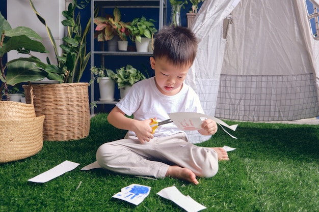 Netter asiatischer Kindergartenjunge, der ein Stück Papier schneidet, Scherenfähigkeiten für Kleinkind einführen