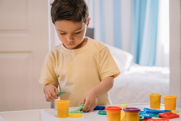 Netter aufmerksamer Junge, der Stock hält, während er mit buntem Plastilin modelliert