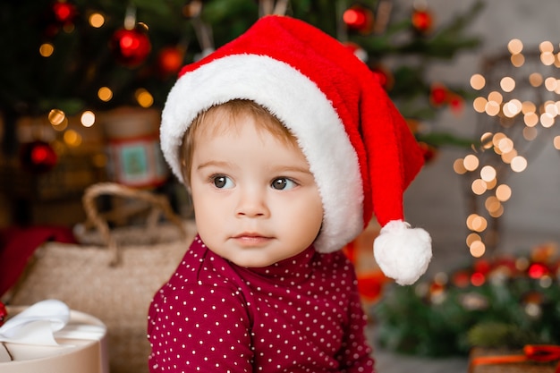 Netter Baby-Weihnachtsmann sitzt zu Hause nahe dem Weihnachtsbaum mit Geschenken