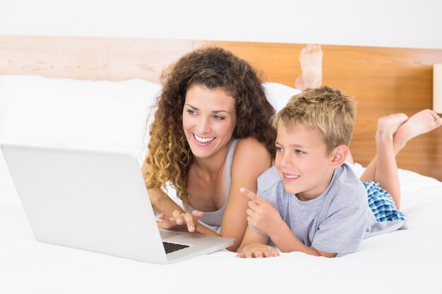 Netter blonder Junge und Mutter, die auf Bett unter Verwendung des Laptops liegt