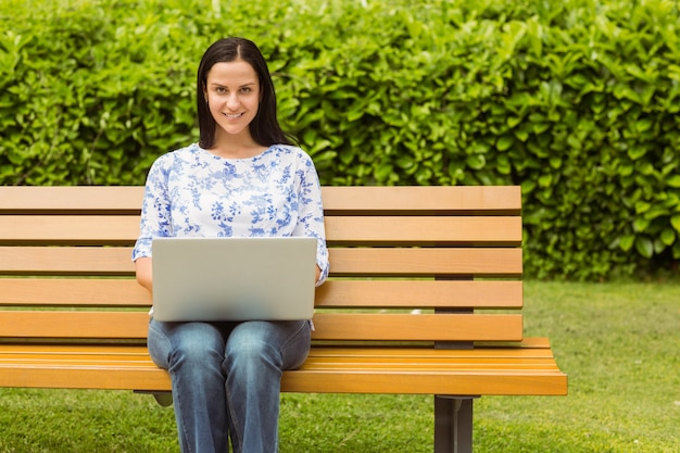 Netter Brunette, der auf Bank unter Verwendung des Laptops sitzt