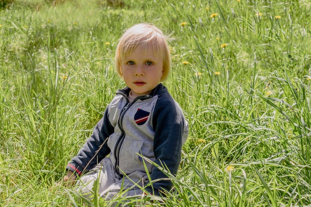 Netter entzückender kleiner kaukasischer Kleinkindjunge, der Spaß hat, auf grünem Grasrasen zu Hause im Hof zu sitzen