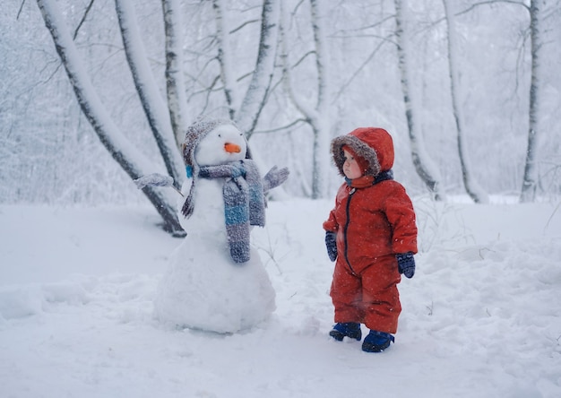 Netter europäischer Junge und der lustige Schneemann in den verschneiten Wäldern an einem Wintertag im Schnee