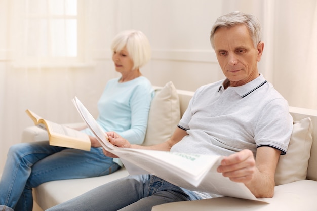 Netter fokussierter älterer Mann, der in einem Wohnzimmer sitzt und Morgenzeitung genießt, während seine Frau ein Buch liest