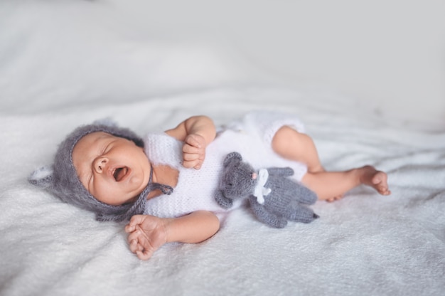 Netter gähnender neugeborener Säuglingsbaby mit Spielzeug, das im Kinderbett in einem Strickanzug mit Ohren schläft.