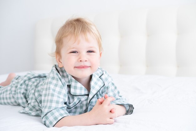 Netter gesunder Kleinkindblonder Junge im karierten Pyjama, der Spaß hat, im weißen Bett zu Hause liegend.