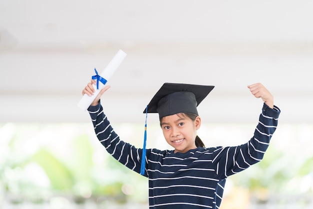 Netter glücklicher asiatischer Schulkindabsolvent mit Abschlusshut und einem Diplom lokalisiert auf weißem Hintergrund