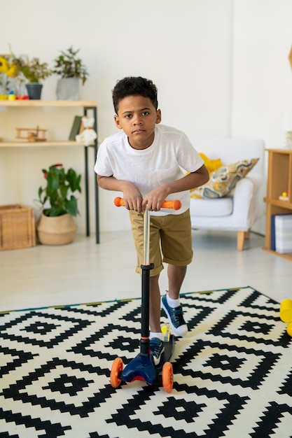 Netter glücklicher gemischter Vorschulkind in der Freizeitkleidung, die Spielzeugplastikroller auf Schwarzweiss-Teppich im Wohnzimmer oder im Kindergarten fährt