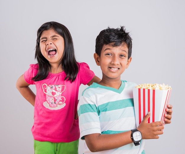 Netter glücklicher kleiner indischer Junge und Mädchen oder Geschwister, die Popcorn essen und fernsehen, während sie auf dem Stuhl sitzen