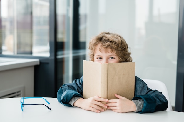 Netter glücklicher kleiner Schuljunge, der aus dem offenen Buch bei der Lektion der Literatur herausschaut, während er am Schreibtisch sitzt