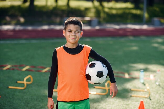 Netter hispanischer Junge in der Fußballuniform mit Fußball nach intensivem Training auf Stadion