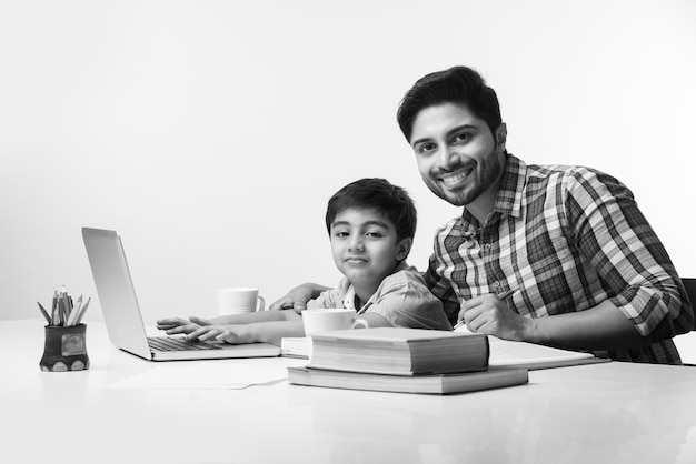 Netter indischer Junge mit Vater oder männlichem Tutor, der zu Hause mit Laptop und Büchern Hausaufgaben macht - Online-Schulungskonzept