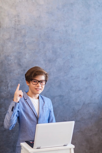 Netter jugendlich Junge mit Finger oben und Laptop gegen Wand