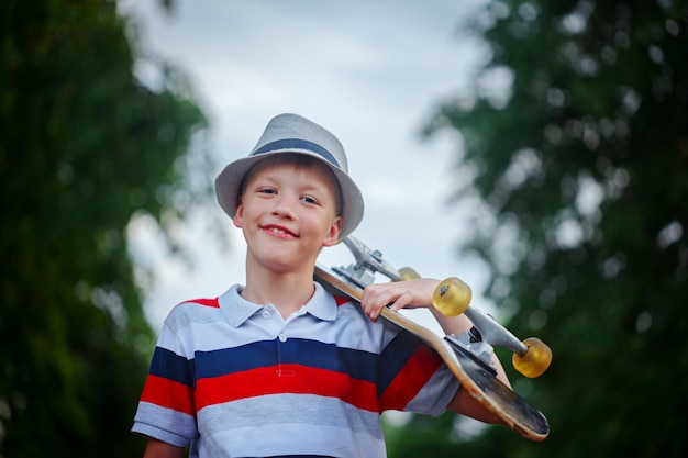 Netter Junge, der draußen Skateboard in der Hand hält Tragende Kappe und stilvolle Kleidung