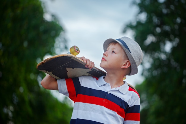 Netter Junge, der draußen Skateboard in der Hand hält Tragende Kappe und stilvolle Kleidung