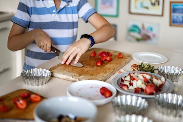 Foto netter junge, der frische beerenerdbeere auf holzbrett schneidet, verwenden messer, das sommerdessert in der küche kocht