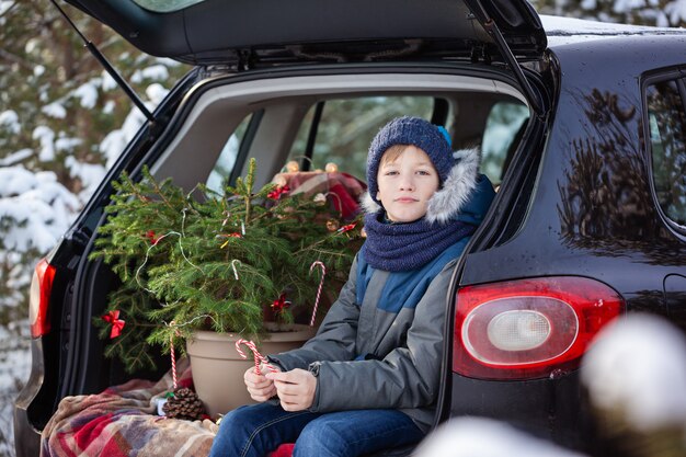 Netter Junge, der im schwarzen Auto am schneebedeckten Winterwald sitzt. Weihnachtskonzept.