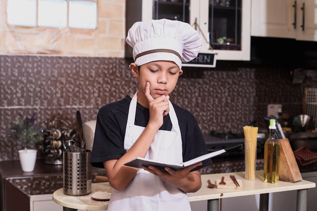 Netter junge, der kochuniform benutzt, denkt, während er ein buchrezept liest, das sich auf das kochen im kitc vorbereitet