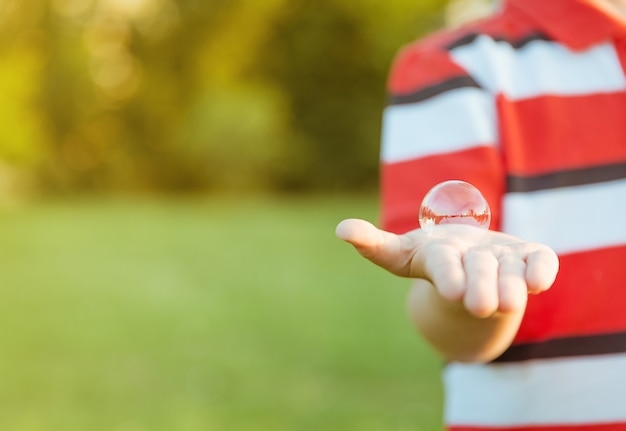 Netter Junge, der Seifenblase in seiner offenen Hand über einem Naturhintergrund zeigt