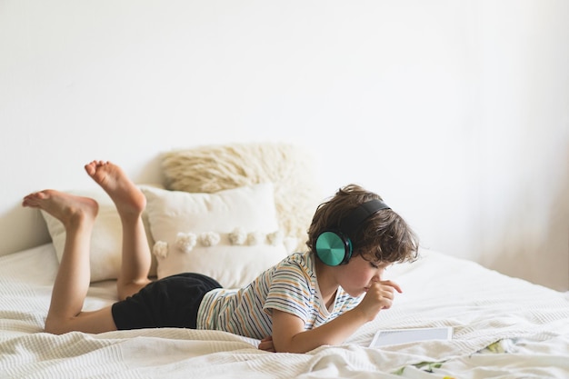 Netter junge, der zu hause auf dem bett liegt und mit pc-tablet spielt oder online liest und musik mit drahtlosen kopfhörern hört