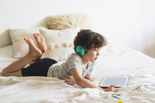 Netter junge, der zu hause auf dem bett liegt und mit pc-tablet spielt oder online liest und musik mit drahtlosen kopfhörern hört