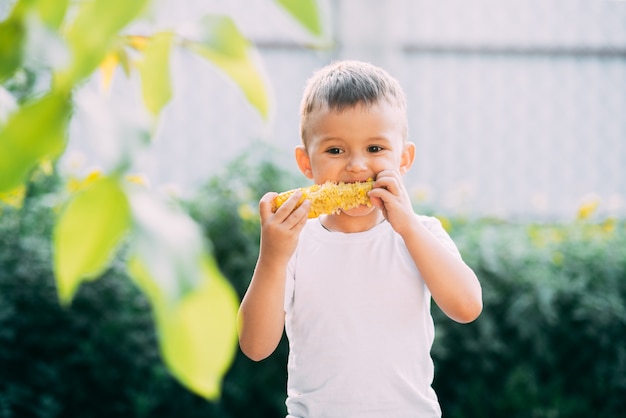 Netter Junge im Garten oder im Freienpark, der gekochten Mais isst, ist im Sommer sehr appetitlich