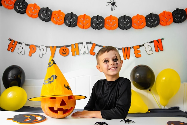 Netter Junge im Kostüm, der hinter dem Tisch im Halloween-Thema dekorierten Raum Halloween von Covid19 sitzt