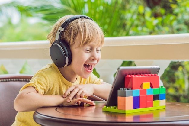 Netter Junge in den Tropen, der mit Freunden und Familie per Videoanruf über ein Tablet und drahtlose Kopfhörer spricht, die auf der Terrasse sitzen.