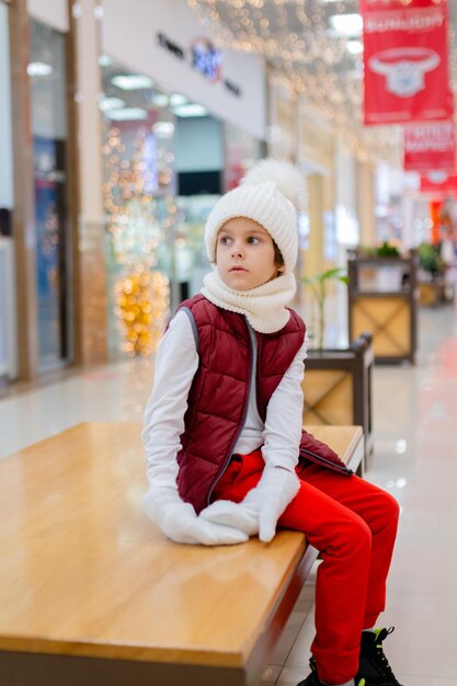 Netter Junge in einem weißen, flauschigen Hut in einem Einkaufszentrum vor dem Hintergrund heller Lichter