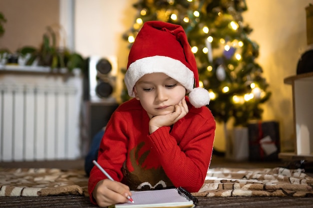 Netter Junge in rotem Pullover und roter Weihnachtsmütze schreibt einen Brief an den Weihnachtsmann und denkt an Geschenke