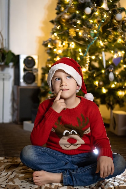 Netter Junge in rotem Pullover und roter Weihnachtsmütze schreibt einen Brief an den Weihnachtsmann und denkt an Geschenke