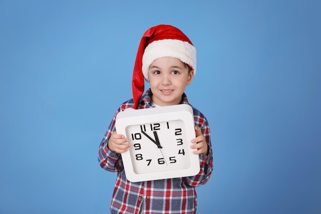 Netter Junge in Weihnachtsmütze mit Uhr auf Farbhintergrund. Weihnachts-Countdown-Konzept