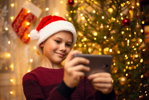 Netter Junge macht ein Selfie im Hut des Weihnachtsmanns, geschmückter Tannenbaum. Das Konzept von Weihnachten.