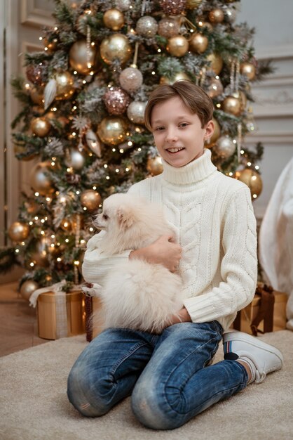 Netter Junge mit einem Hund in den Armen in einem leichten Pullover auf dem Hintergrund eines Weihnachtsbaumes