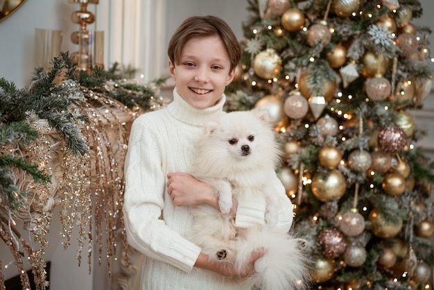 Netter Junge mit einem Hund in den Armen in einem leichten Pullover auf dem Hintergrund eines Weihnachtsbaumes