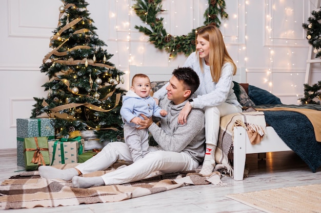 Netter Junge mit glücklichen Eltern in der Nähe des Weihnachtsbaums Eine Familie mit einem kleinen Kind in einem Schlafzimmer in der Nähe eines geschmückten Weihnachtsbaums Festliche Neujahrsatmosphäre Familienfeier