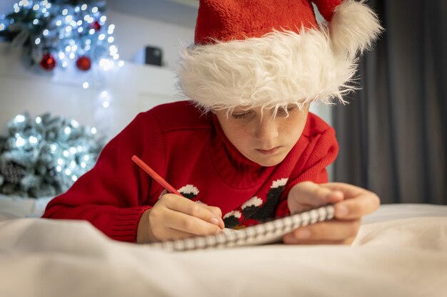 Netter Junge schreibt Brief an den Weihnachtsmann. Vorbereitung auf den Urlaub. Weihnachtswunder-Wunschliste. Kind, das zu Hause mit Weihnachtslicht auf dem Bett liegt. Weihnachtsstimmung, Feiertagsfeiern Konzept hautnah.