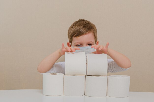 Netter Junge von vier in einer schützenden medizinischen Maske spielt mit Toilettenpapier