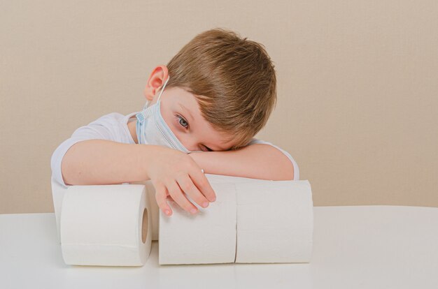 Netter Junge von vier in einer schützenden medizinischen Maske spielt mit Toilettenpapier