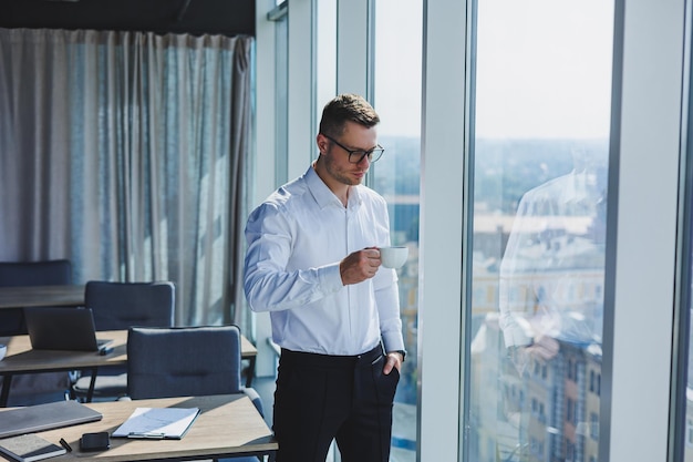 Netter junger Geschäftsmann mit Kaffee in der Hand in Gläsern und weißem Hemd ist er in einem modernen Büro mit großen Fenstern Ein Mann in klassischen Hosen und einem weißen Hemd Junger attraktiver Geschäftsmann