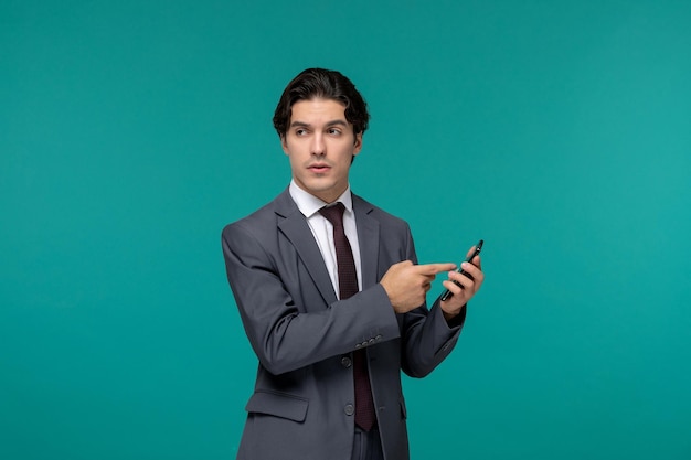 Netter junger gutaussehender mann des geschäftsmannes im grauen büroanzug und -krawatte, die auf telefon zeigen