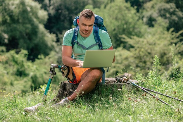 Netter junger männlicher Tourist, der auf dem Baumstumpf sitzt, während er seinen Laptop benutzt