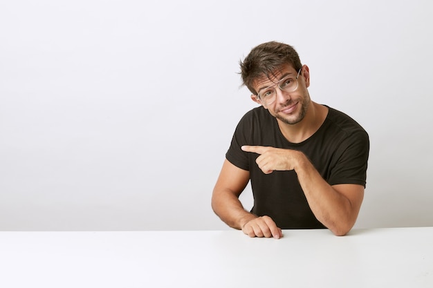 Netter junger Mann mit Brille, die auf weißem Schreibtisch zur Seite zeigt und sich auf weißen Schreibtisch stützt. Schwarzes T-Shirt tragen. Produkt präsentieren.